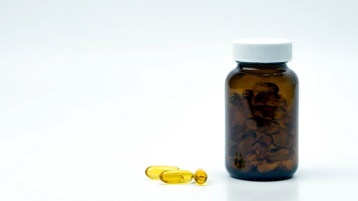 A clear glass bottle filled with small beige capsules standing on a light wooden shelf.